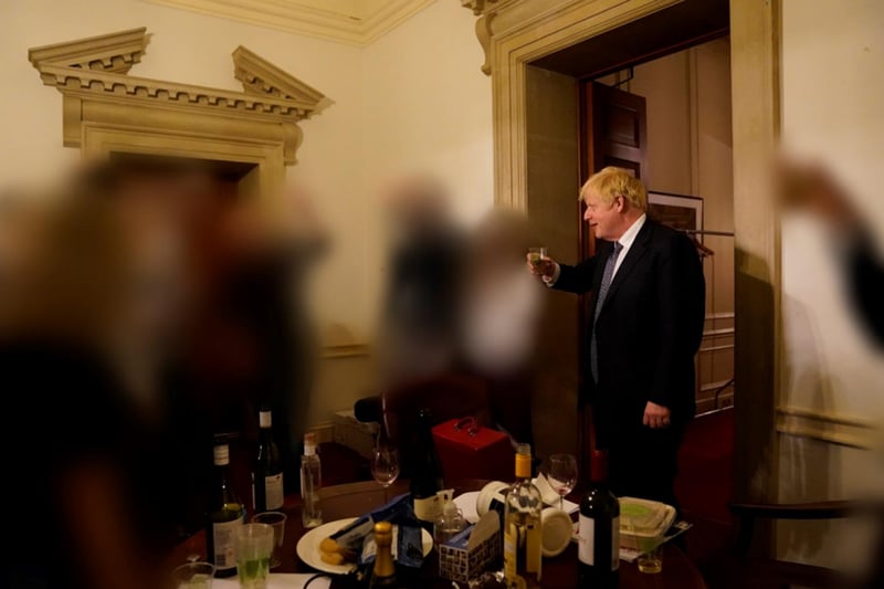 The PM chats while holding his drink. Several bottles of wine are visible on the table in front of him.