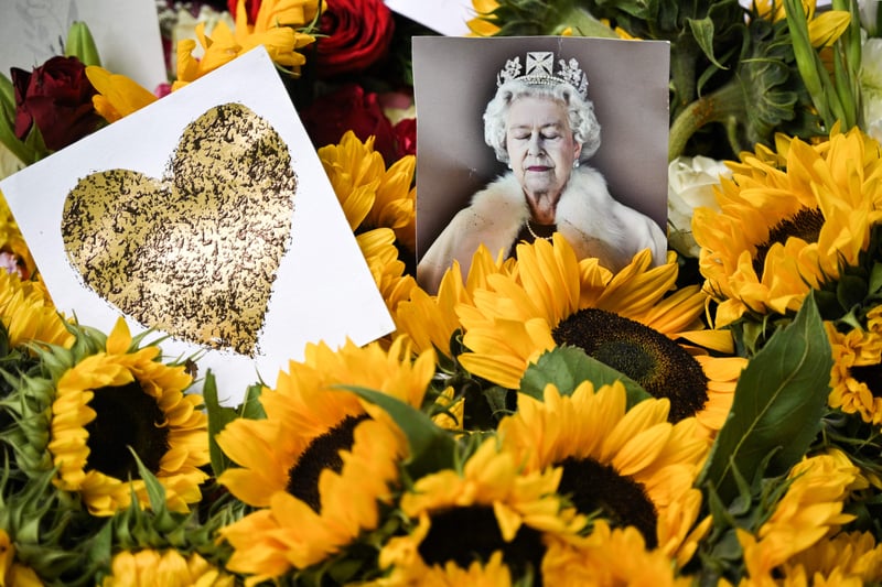 The older Queen dressed in white with her eyes closed, next to a golden heart amid a display of yellow sunflowers.