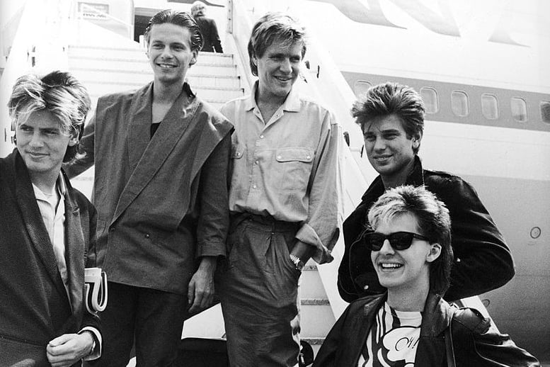 Duran Duran smile and laugh while posing on an airplane ramp before departing for Australia, Heathrow Airport, London, England, July 26, 1983 (Photo by Express Newspapers/Getty Images)