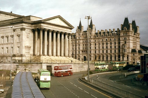 Red and green buses drive past St George’s Hall. The building on the other side of Lime Street Station was originally the North Western Hotel and the station is immediately behind it.