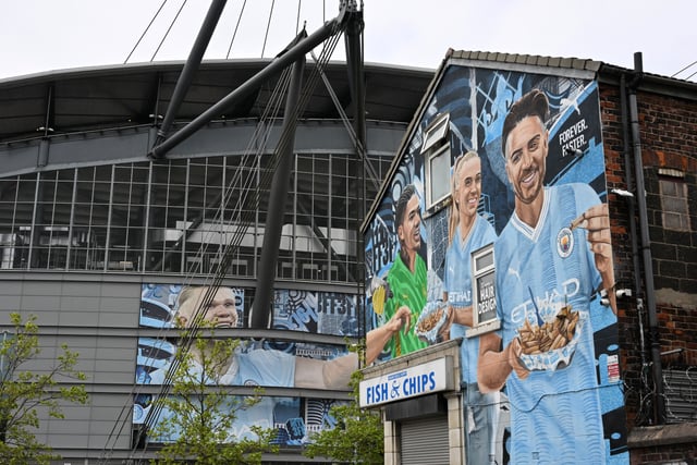 Street art murals, one on the wall of a Fish and Chip shop (R) and one on the outside of the Etihad Stadium (L), depict Manchester City player Norwegian striker Erling Haaland (L) taking a chip from Brazilian goalkeeper Ederson (C) and English defender Esme Morgan (2R), along with Manchester City's English midfielder Jack Grealish eating chips