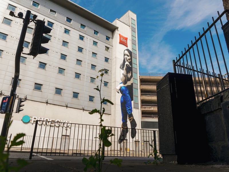 Glasgow artist James Klinge made his Nuart debut in style in 2022, with his art beginning with detailed hand cut stencils and descending into self-described “controlled chaos”. The chaos Klinge describes is likely contained to his process, as his finished artwork sits above a busy road with a woman cheekily calling for silence. Though his portrait perches on the wall of the Ibis Hotel, absolute silence isn’t likely with its location along the A956 Virginia Street and opposite the harbour.