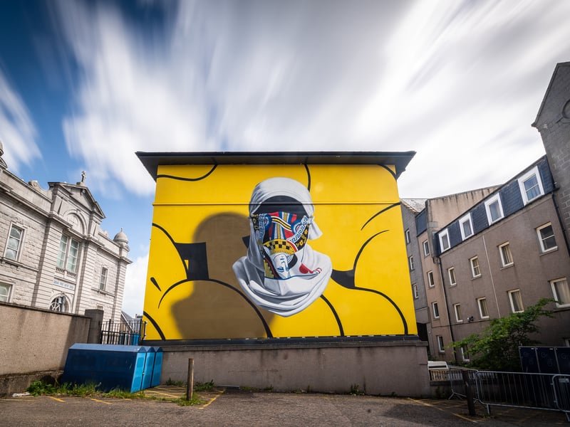 Nuno Viegas, also known as Metis, is a Portuguese artist inspired by the graffiti scene. Often using his art to present contrast between the visually aggressive reality of traditional graffiti and a peaceful and clean representation of the work, Viegas’ 2022 mural is a bold demonstration of this. Featuring a floating mask and headscarf and surrounded by a vibrant yellow background, the mural is similar to another which Viegas created for the Stavanger festival. Perched on the opposite side of the building to Henrik Uldalen’s 2021 mural which can also be spotted from Gerrard Street, this bold mural brightens up the space around it.