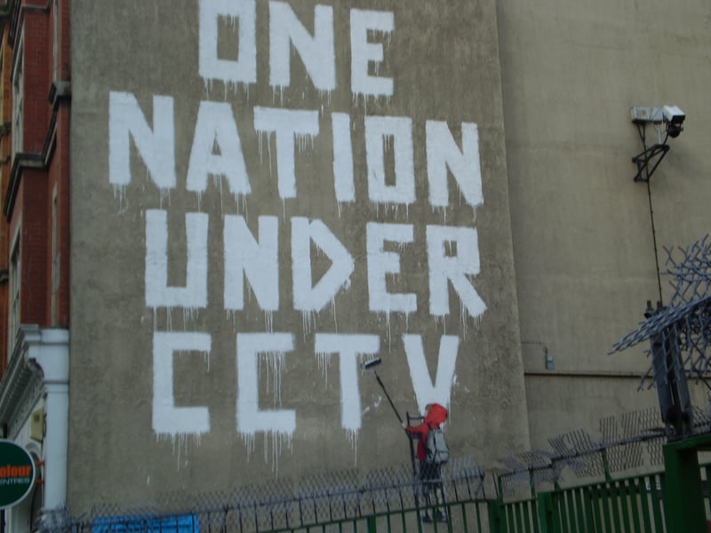 ‘One Nation Under CCTV’ first appeared in Newman Street, London. It depicts a child in a red hood painting “one nation under CCTV” while being supervised by a police officer. The art sits adjacent to a CCTV camera. It poses a warning to the public about the emerging surveillance state of the world. In places like China, the IHS Markit estimated that the country had 540 million cameras in it i.e., more than one camera per three people.