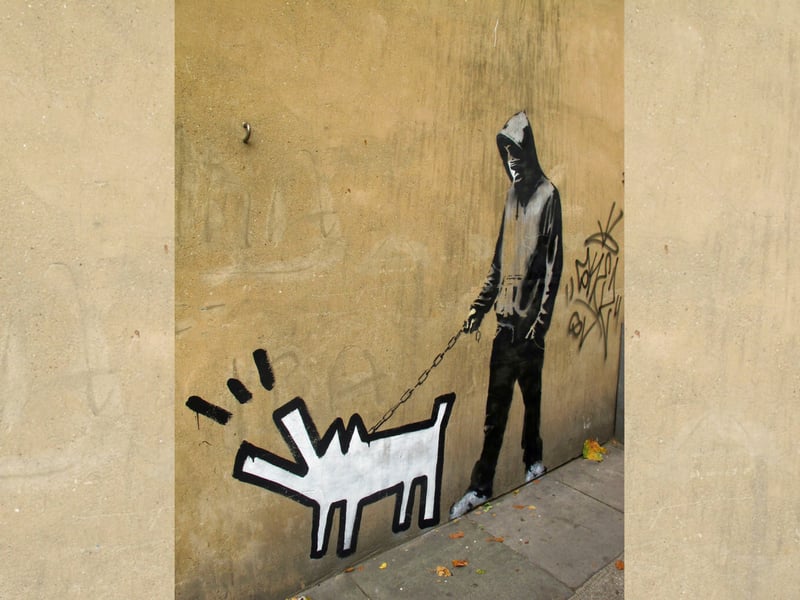 This work of Banksy’s first appeared in the Southwark District of London. It depicts an aloof masked youth with one hand casually stuck in his pocket while the other pulls on a frenzied dog. The Banksy Explained website calls it “a portrayal of British disaffected youth and gang culture whose aggressive dogs have become weaponry in their quest for power.”