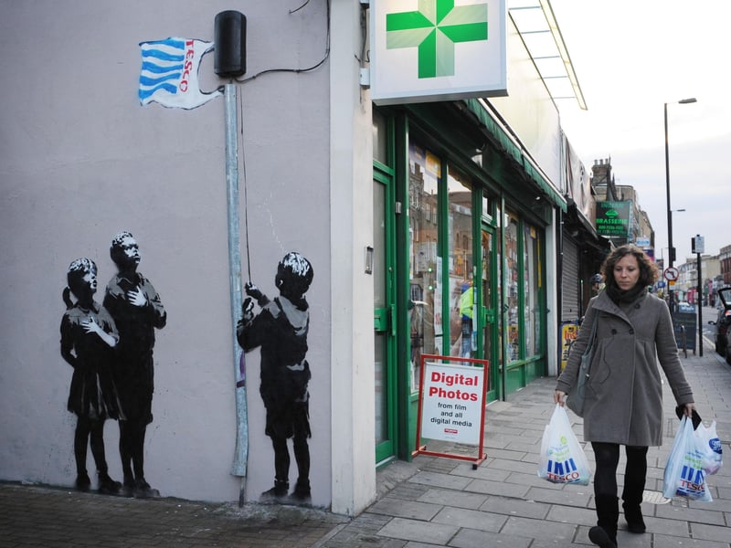 ‘Very Little Helps’ was created next to a pharmacy on Essex Road in North London. It shows a group of children hoisting a Tesco bag as a flag as they gather around the flagpole. The graffiti art is said to be Banksy’s “critique” of the spread of mega-corporations worldwide which is harmful to local (and often rural) communities.