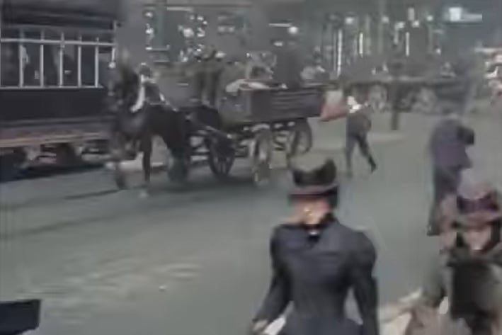 Two well-dressed Victorian woman walk hurriedly down Jamaica Street as a tram passes in the background
