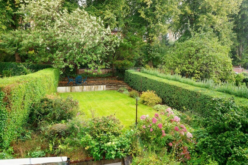 Steps lead up to the well maintained private rear garden with raised patio, mowed lawn area and surrounding beds, enclosed by hedging and boundary wall at rear.