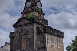 The clock tower is all that remains of the former Pollokshaws Burgh Halls. Erected in 1803 to administer the affairs of the town and to provide a place for meetings, it took ten years to complete. It included a distinctive clock tower much to the envy of neighbouring councils. It covered about half the area of the square, and over the years extensions were added with shops located externally in parts of the old building. One of them was occupied by McClurg’s the Fishmonger who until recently operated a business in The Stag Inn building.  According to Fowlers Commercial Directory for Renfrewshire (1834-35) the licensee was James Struthers. It was partly demolished in 1895 as a result of the opening of the new burgh hall (see No.1) and the main part was demolished in 1934 leaving only the squat tower containing the vestibule. It is still affectionately known by locals as The Toonhoose. 