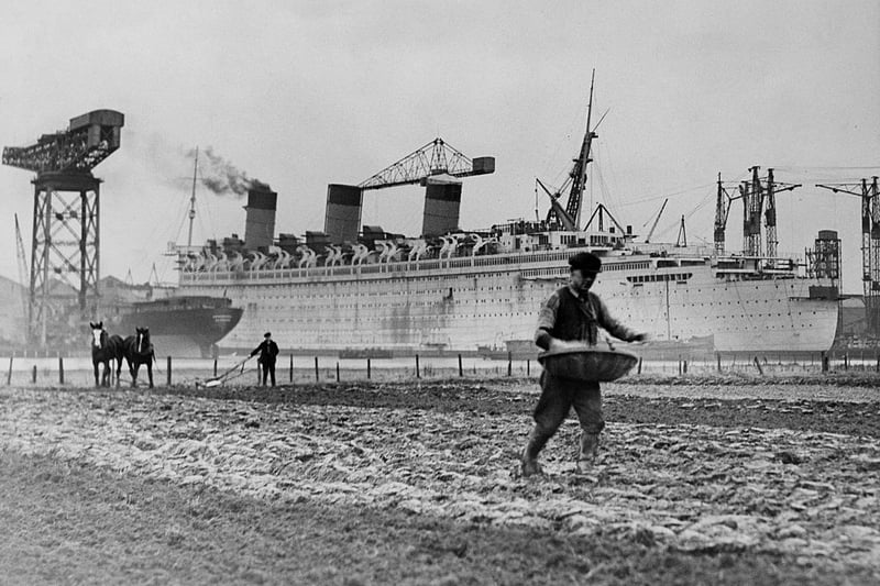 Farmers tilling the soil with horse drawn ploughs and hand sowing their seeds beside the British passenger liner for the Cunard-White Star Line, RMS Queen Mary as she is being built at the John Brown & Company shipyard at Clydebank on 13 January 1936 at Clydebank