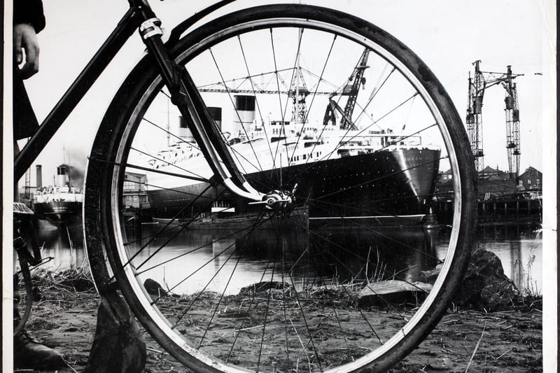 The ocean liner 'RMS Queen Mary' in the fitting-out yard on the river, as seen through the wheel of a bicycle, circa 1935.