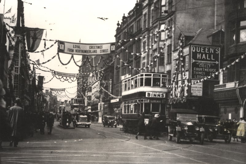  A view of Northumberland Street Newcastle upon Tyne taken in 1935. The photograph shows Northumberland Street decorated for the Silver Jubilee of King George V and Queen Mary. Pearl Assurance Buildings are in the foreground to the right then the entrance to Northumberland Place with Amos Atkinson boot and shoe makers beyond. There is an advert for the Queen’s Hall Northumberland Place on the side wall of Amos Atkinson. Trams buses and cars are travelling down Northumberland Street. A B-class tram is on route 15 and carries an advert for Binns Department Store.  (Newcastle Libraries)