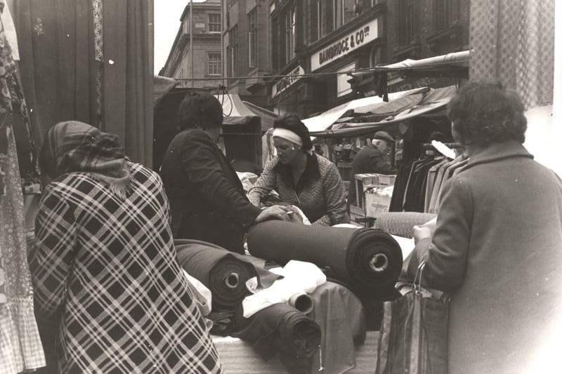 A photograph of the market on the Bigg Market taken c.1970. In the foreground is a stall selling clothes and fabric. Two women are looking at the fabric and a third is making a purchase from the stall owner. Other stalls can be seen in the background. To the right of the stalls is the premises of Bainbridge ( L. Torday)