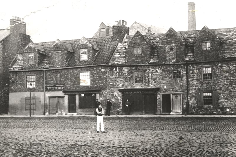 : A view of ‘old houses’ on Barras Bridge taken in 1880. The houses are being used as shops and include a cooper and a carver/gilder. Three men are standing on the pavement in front of the houses and a fourth man is standing in the middle of the cobbled road in the foreground. (Newcastle Libraries)
