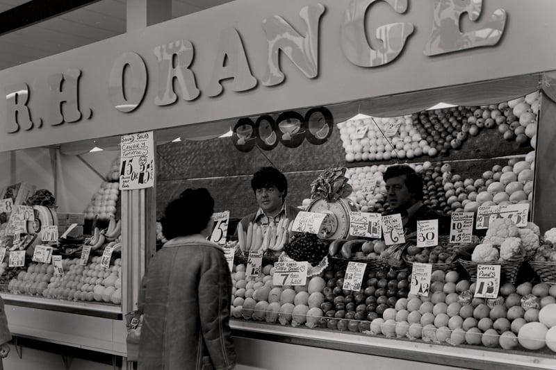 A view of the Green Market Eldon Square Shopping Centre Newcastle upon Tyne taken in 1976. The photograph shows the fruit and vegetable stall of ‘R.H. Orange’. Two members of staff are serving a customer. (Newcastle Libraries)