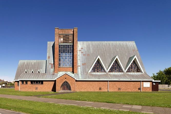 This impressive church is a rare example in the north of England by Lawrence King, one of the leading ecclesiastical architects of the post-war period. Built between 1960 and 1962, its bold sculptural design in the form of an upturned boat is dominated by sheer tower walls and tall copper clad roofs with unusual triangular dormer windows representing sails. Its upturned boat design was created by King to emphasise Fleetwood’s strong maritime connections, and its dedication to St Nicholas, the Patron Saint of Sailors, cements this association.
The light and lofty interior, with multiple trusses rising from the ground like the ribs of a ship, even incorporates red and green port and starboard lights either side of the crossing. There are many original fixtures and fittings including some very finely carved and painted statues of The Virgin Mary and St Nicholas, designed by the architect himself. Lawrence King was a gifted designer and an important voice in the addition of artworks to churches after the Second World War (Faith Craft) which produced different works intended for the beautification of worship.