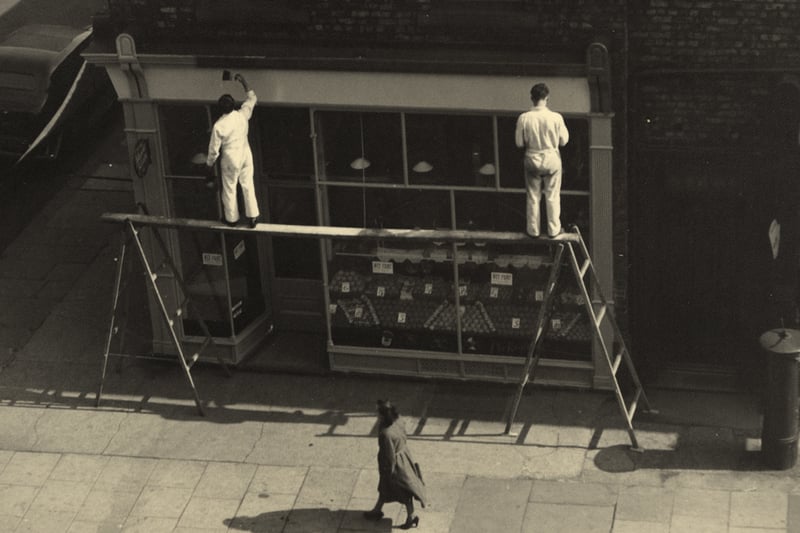 A view of Sandyford Road/Marianople Street Jesmond taken in the 1950s. The photograph shows two workmen standing on a wooden plank supported by two ladders painting the woodwork around a shop window.