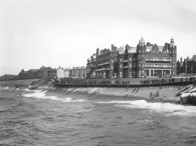 The Metropole Hotel, 1890-1910. The north shore with people walking along the promenade by the Victorian Hotel Metropole. Beach huts have been pulled closer to the railings out of the way of the rough sea