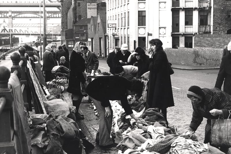 A view of Paddy's Market Quayside Newcastle upon Tyne taken in 1975. The photograph shows clothes for sale on railings tables and on the ground. Vendors and customers are standing beside the clothes. The Tyne Swing and High Level Bridges can be seen in the background.