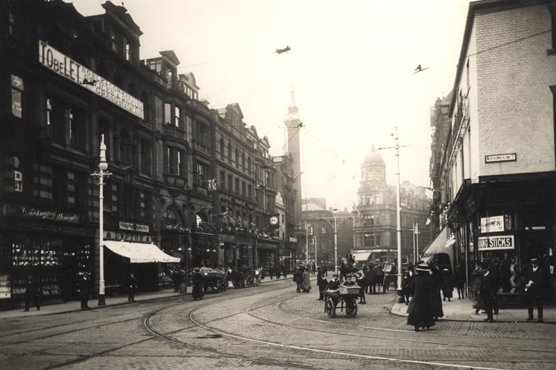 A view of Blackett Street decorated for the coronation of King George V and Queen Mary in 1911. It is a busy street scene with a variety of horse-drawn vehicles travelling along the road. In the foreground two boys are pushing a cart. The buildings on the left include 'Northern Goldsmiths' and 'Lipton'. Grey's Monument and the YMCA building can be seen in the distance. Tramlines and wires can be seen in the foreground.
