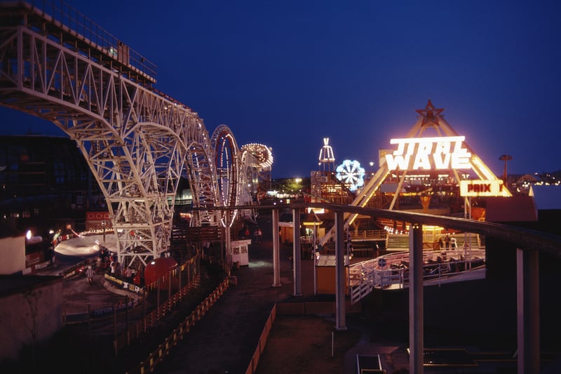 A night view of the illuminated rollercoaster and fun rides including the 'Tidal Wave' at the seaside resort of Blackpool, 1983