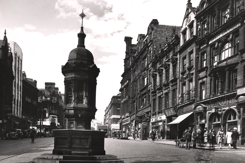 A photograph of the Bigg Market taken c.1947. In the foreground is the memorial fountain to Dr J. H. Rutherford a temporance reformer. On the left-hand side of the Bigg Market there are buildings containing offices and shops such as the Roma Cafe and on the right-hand side buildings include the Half Moon Inn and Bainbridge and Co. Ltd. The premises of H. Samuel jewellers can be seen in the background on the corner of Grainger Street and Newgate Street. 