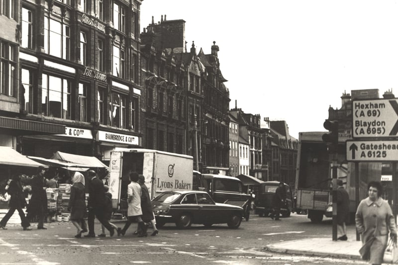 A photograph of the Bigg Market on market day taken c.1972. The view is looking across to the left-hand side of the Bigg Market. People are crossing the road in the foreground. The left-hand side of the road is lined with stalls and vans are parked behind them.