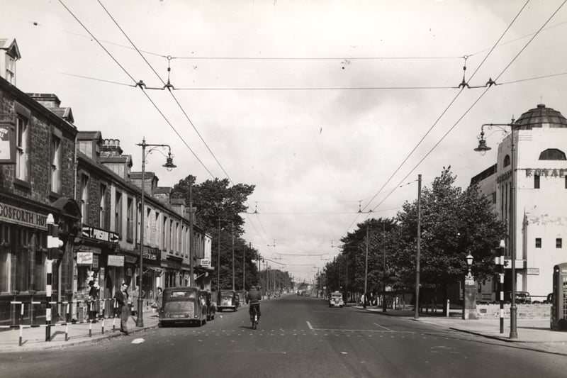 A view of the High Street Gosforth taken c.1960. The photograph is looking north and shows buildings on both sides of the High Street. The Gosforth Hotel is in the foreground to the left with shops beyond. The Royalty cinema is in the centre to the right with trees in front. Several cars are parked in front of the shops on the left-hand side of the street and a cyclist and cars can be seen travelling along the road. 