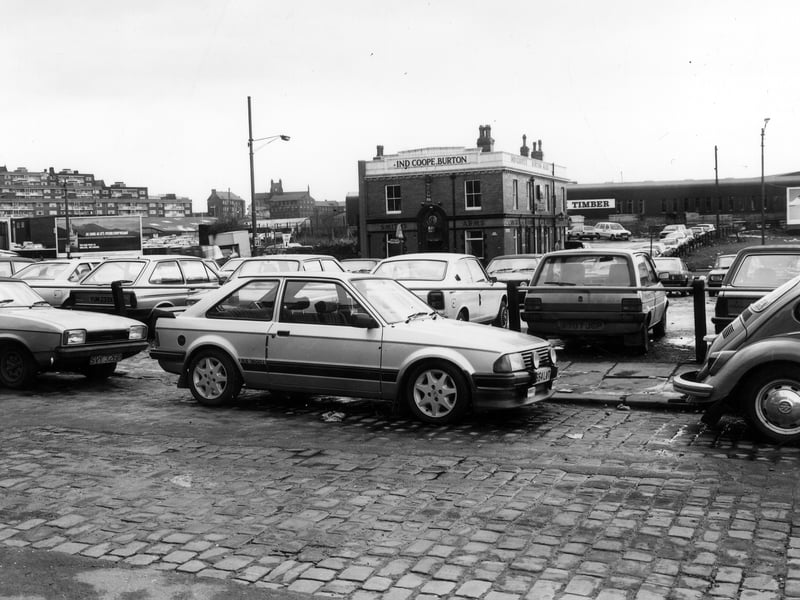 A view looking from Woolman Street across a busy car park towards Marsh Lane in January 1985. Cars are also parked at the side of Woolman Street in the foreground. The Smith's Arms public house is seen in the centre background, standing alone as buildings adjoining it on the left have recently been demolished. Rising up on the far left are the flats of Saxton Gardens.