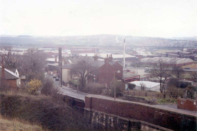 Millshaw looking south-west with the railway bridge in the foreground on the right. Old buildings, including Holly House in the centre, are located on the roadside while further back are newer industrial buildings. Cottingley Housing Estate is seen in the background on the far right with Churwell Viaduct behind and Churwell beyond this.