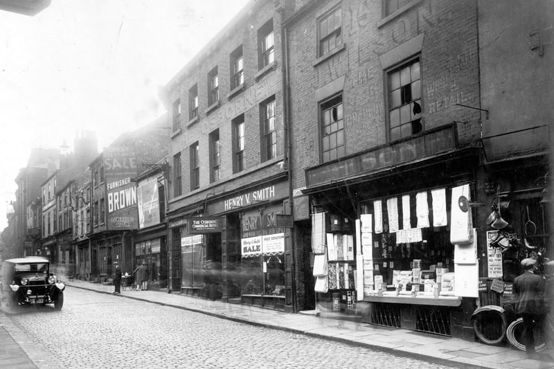 Lower Briggate in September 1928. September 1928 View looking north west, prior to widening and redevelopment of The Headrow. From right, number 17 Mrs Annie Lewis, cycle dealer, there are bicycle parts hanging outside the shop. Next, number 16 John Wilson draper with materials in window and outside. Number 15, Henry V. Smith motor engineer. Entrance to Camden Court with sign for the Osborne Commercial Hotel. Number 14 Albert Doughty, dress materials. Brown and co house furnishers are at 12/13.