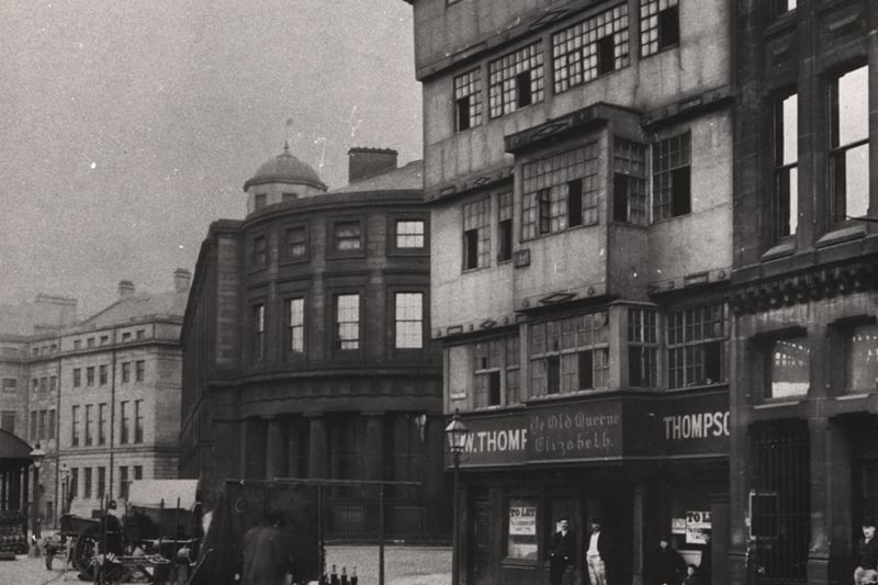 The corner of the Quayside. In the back centre of the picture stands the Guild Hall. The corner building on the right of the picture is slightly run-down with missing windows. The lower floor is being advertised as 'To Let' as well as bearing the painted signs 'Ye Olde Queen Elizabeth' and 'W. Thompson'. Onlookers crowd the entrance. A market vendor stands with his merchandise (some form of bottled liquid) in the middle of the street. 