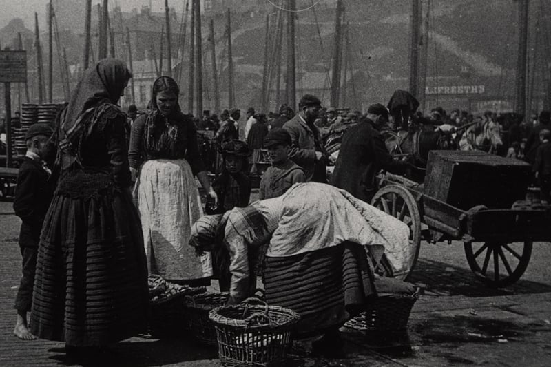 A photograph of a group of three Fishwives sorting herring into baskets North Shields c.1898. The Fishwives are wearing their traditional working clothes which include a jacket or bedgown a shawl an ankle or mid-calf length skirt with tucked hemline and an apron. A group of children are watching the women as they work. In the background the Quayside is busy with people.