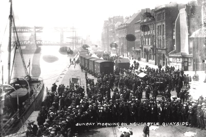 A view of the Quayside Market Newcastle upon Tyne taken c.1900. The photograph shows the market on a busy Sunday. A large crowd has gathered in the foreground with a sailing ship moored on the Quayside to the left. There is a row of goods vans in the centre of the market and a second sailing ship moored further up the quay