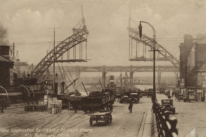 The photograph shows the bridge being built the two sides of the arch are being held in place by cables waiting for the middle section to be inserted. The photograph is taken from Newcastle quayside which is busy with cars and lorries.