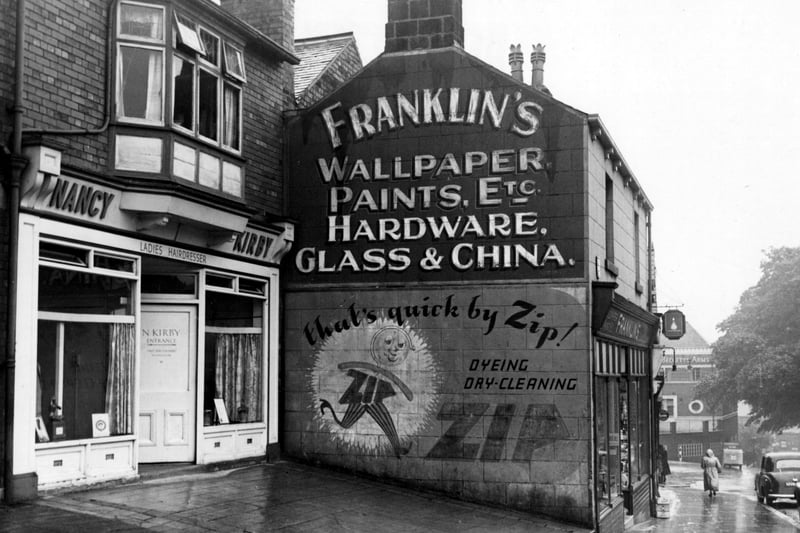 Shops on Stonegate Road in August 1953. Nancy Kirby ladies hairdresser and Franklin's wallpaper, paints, etc. shown. Beckett Arms pub is on the corner with Meanwood Road in the background. Advertisement for Zip dry cleaning on side of shop.
