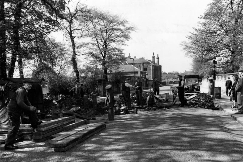 Road subsidence on Stainbeck Lane with maintenance work in progress. A car, a bus stop, workmen and onlookers are visible. Pictured in May 1953.