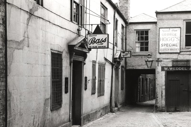  A 1962 photograph of Ye Olde George pub taken from the Cloth Market looking into Old George Yard. The frontage of Ye Olde George can be seen. An upper-storey room links Ye Olde George with a garage which is to the right of the building. Underneath the room there is an entranceway which gives access to the buildings beyond Ye Olde George. The garage is owned by 'W. Weddel.