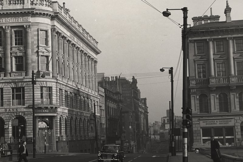 The photograph is taken from Collingwood Street. The Midland Bank is on the corner of the Bigg market and the Newcastle and Gateshead Building Society is on the corner of St. Nicholas' Street. A man is crossing the road by the lights an old man is watching him.