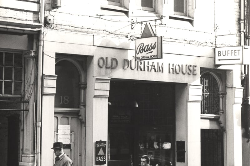 A 1964 photograph of the Old Durham House pub Cloth Market. The view shows the front of the pub and part of the adjoining buildings. In the foreground two men are walking in front of the pub.