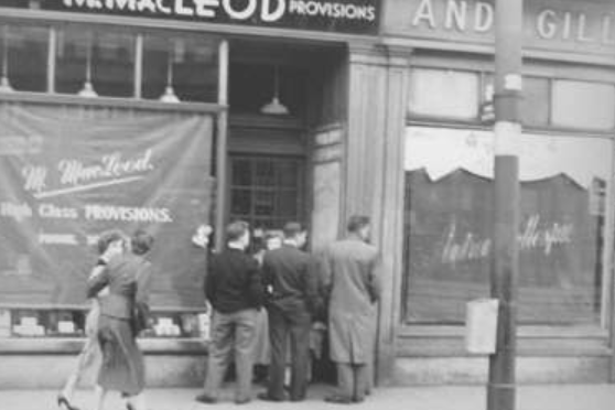 A group of young men crowd round a door outside MacLeod's Grocers on Dumbarton Road