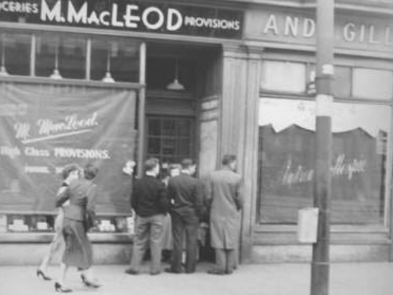 A group of young men crowd round a door outside MacLeod's Grocers on Dumbarton Road