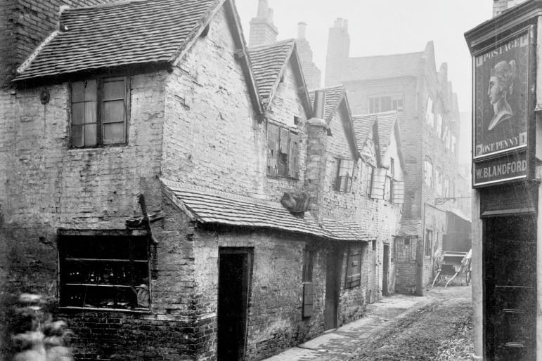 The area is pictured badly run-down. The photograph was possibly taken in the nineteenth century as there is a cart further down the alley and no signs of modernisation. The ground is cobbled with no tarmac visible anywhere. The Queen's head appears in the 'penny black' sign above the doorway to a public house on the right hand side. (Photo by English Heritage/Heritage Images/Getty Images)