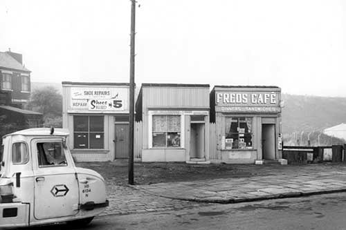 Cow Close Road in October 1969. In focus is a cobblers, offering 'Shoes heeled in 5 minutes'. In the centre is Elizabeth hair stylist with an advert for Ladyset perms in the window. This is number 45. On the right is number 47, Fred's Cafe offering dinners and sandwiches. A crate of milk bottles can be seen to the left of the doorstep. In the foreground on the left edge is part of a three wheeled vehicle with a distinctive logo on the door.