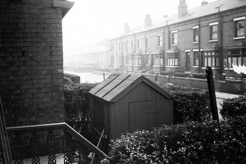Houses on the south side of Cross Flatts Parade from the front of number 15. In the foreground are the railings, wall, hedge and garden shed of Number 15. Terraced houses over the road lead to the junction of Wooler Road. Pictured in October 1952.