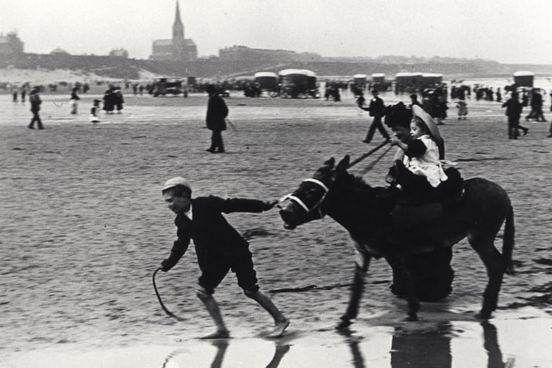 A view of Tynemouth sands taken c.1898. In the foreground a small child is sitting on a donkey which is being pulled by an older boy. A woman is walking beside the donkey. Bathing machines can be seen at the water's edge and parked on the sands.