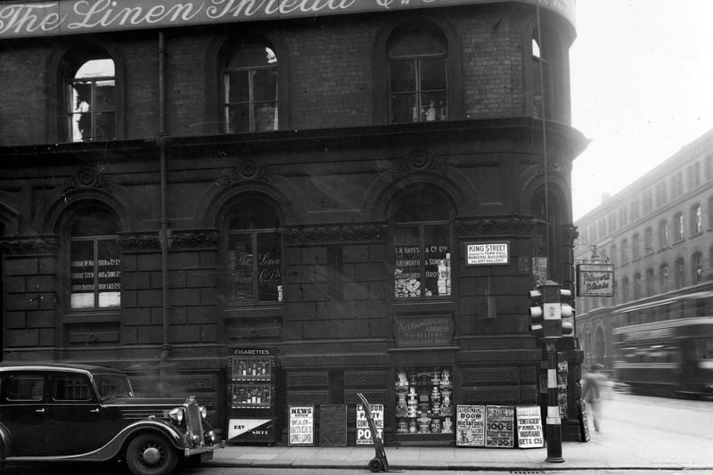 King Street and Wellington Street junction in June 1939, showing The Linen Thread Co. Ltd., F.W.Hayes and Co. Ltd., Ainsworth and Sons Ltd., Crawford Bros. Ltd., Dunbar McMaster & Co.Ltd., Robert Stewart & Sons Ltd., North British Boot Co. Ltd. Also shown are a car, tram, cigarette machine and a porter's trolley. The advertisements are mainly for newspapers with headlines eg. War-Hitler's confession. 