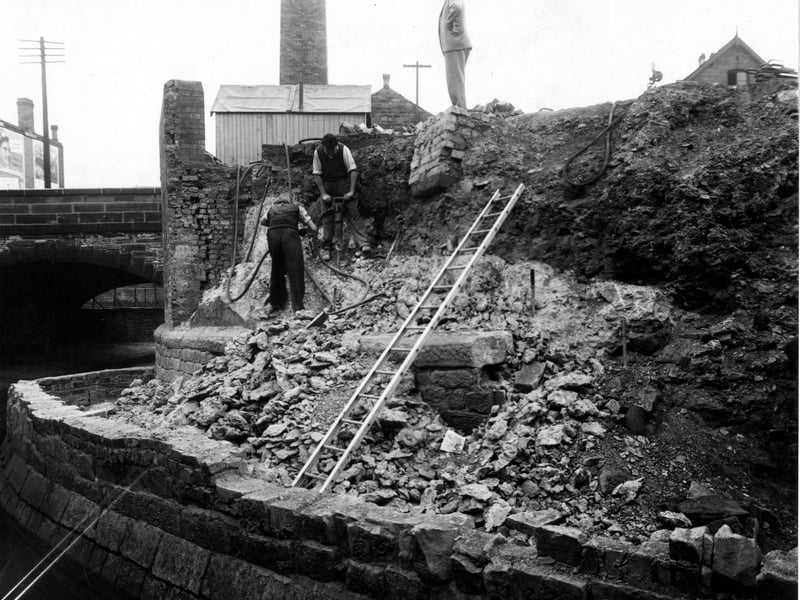 Work on a new retaining wall for Sheepscar Beck near Sackville Street. Some workmen stand on the banking, one is using a pneumatic drill. An arched bridge is to the left, with a workman's hut on top. A ladder is in the foreground.. Pictured in September 1949.