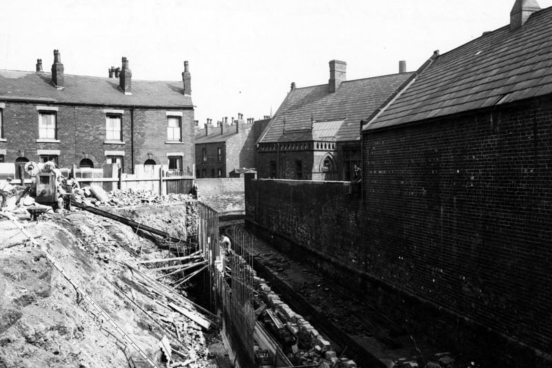 Work in progress on Sheepscar Beck's retaining wall near Sackville Street. Some brick, terraced houses are behind. Metal supports are at the base of the bank, with a cement mixer and some workmen at the top. A man sits on a high brick wall on the right. Pictured in August 1949.