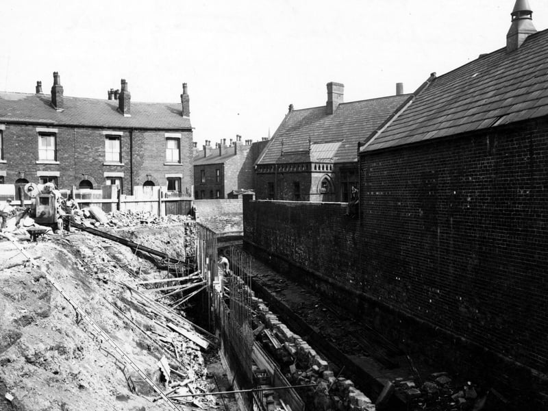 Work in progress on Sheepscar Beck's retaining wall near Sackville Street. Some brick, terraced houses are behind. Metal supports are at the base of the bank, with a cement mixer and some workmen at the top. A man sits on a high brick wall on the right. Pictured in August 1949.